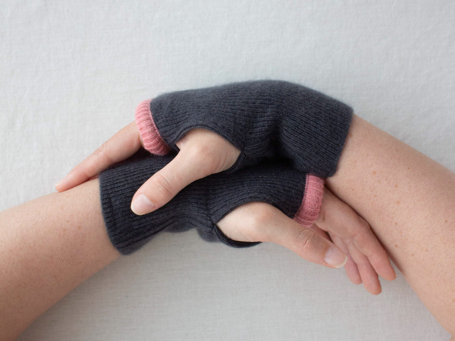 Person wearing dark grey cashmere fingerless gloves with pink cuffs on a light grey background