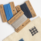 Five spools of yarn in blue, navy, and light beige, each mounted on a brown kraft paper label, arranged on a white surface above a cream woven fabric swatch with small geometric color samples.


