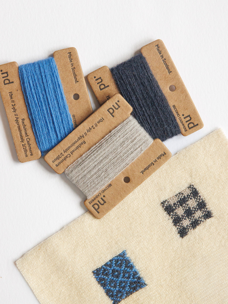 Five spools of yarn in blue, navy, and light beige, each mounted on a brown kraft paper label, arranged on a white surface above a cream woven fabric swatch with small geometric color samples.