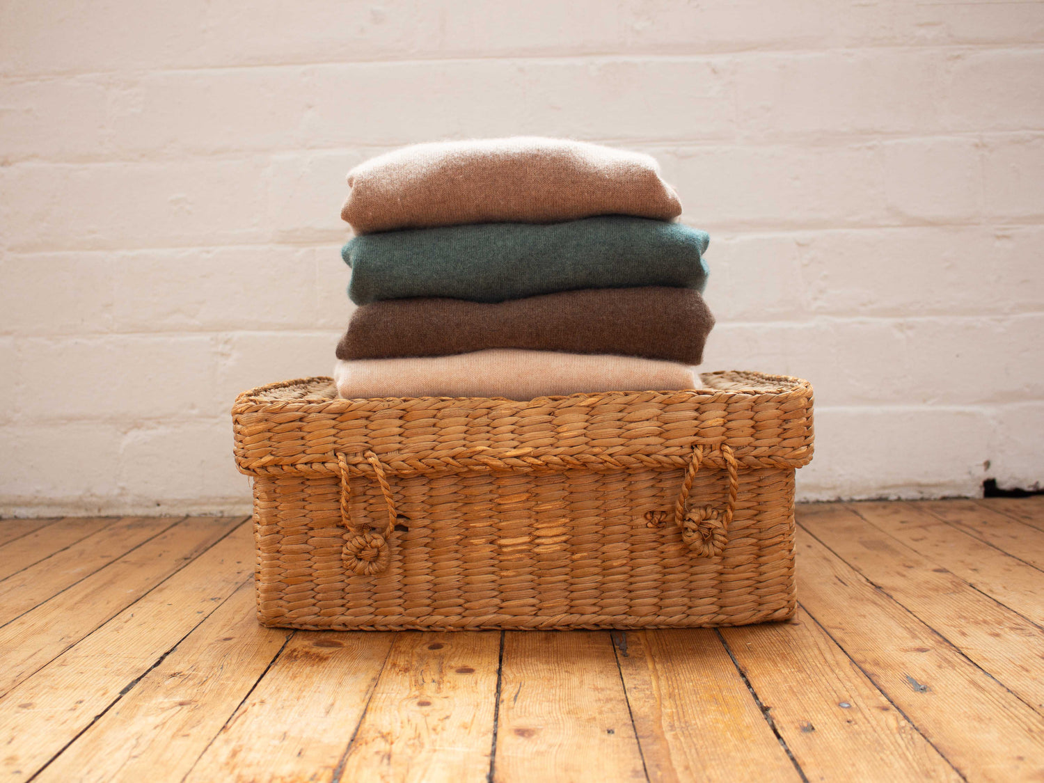 Wicker basket with folded cashmere on a wooden floor against a white brick wall