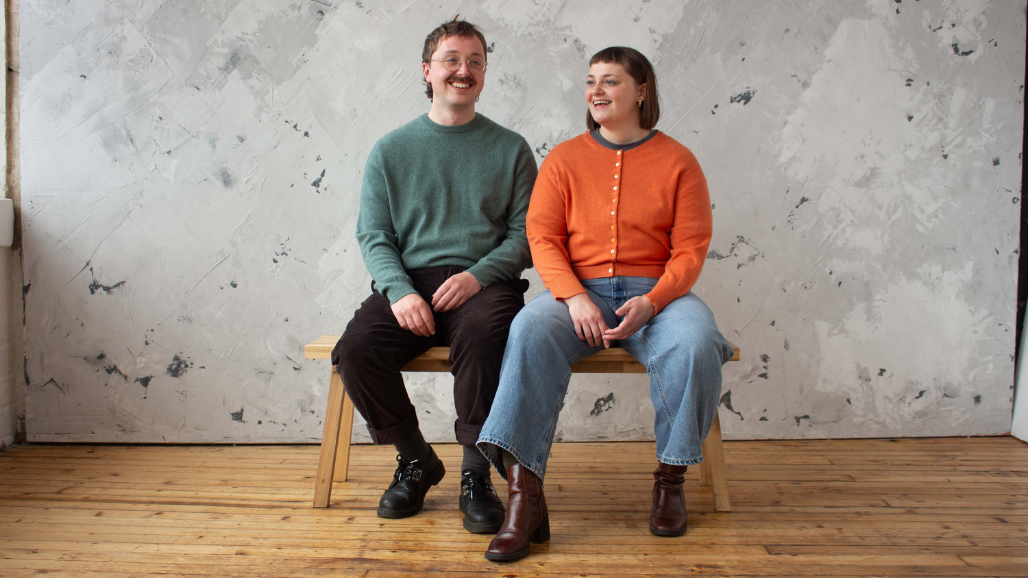 Two people sitting on a wooden stool against a textured wall.